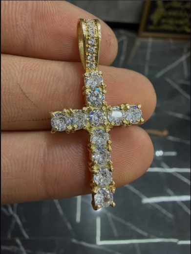 Pingente dourado de cruz com pedras transparentes, segurado para mostrar os detalhes
